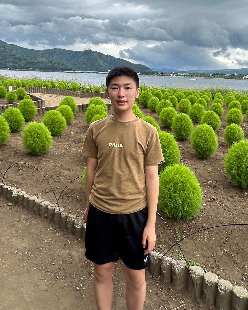 Grant Zhong standing on a path between rows of kochia bushes, with a lake and clouded hills behind him.